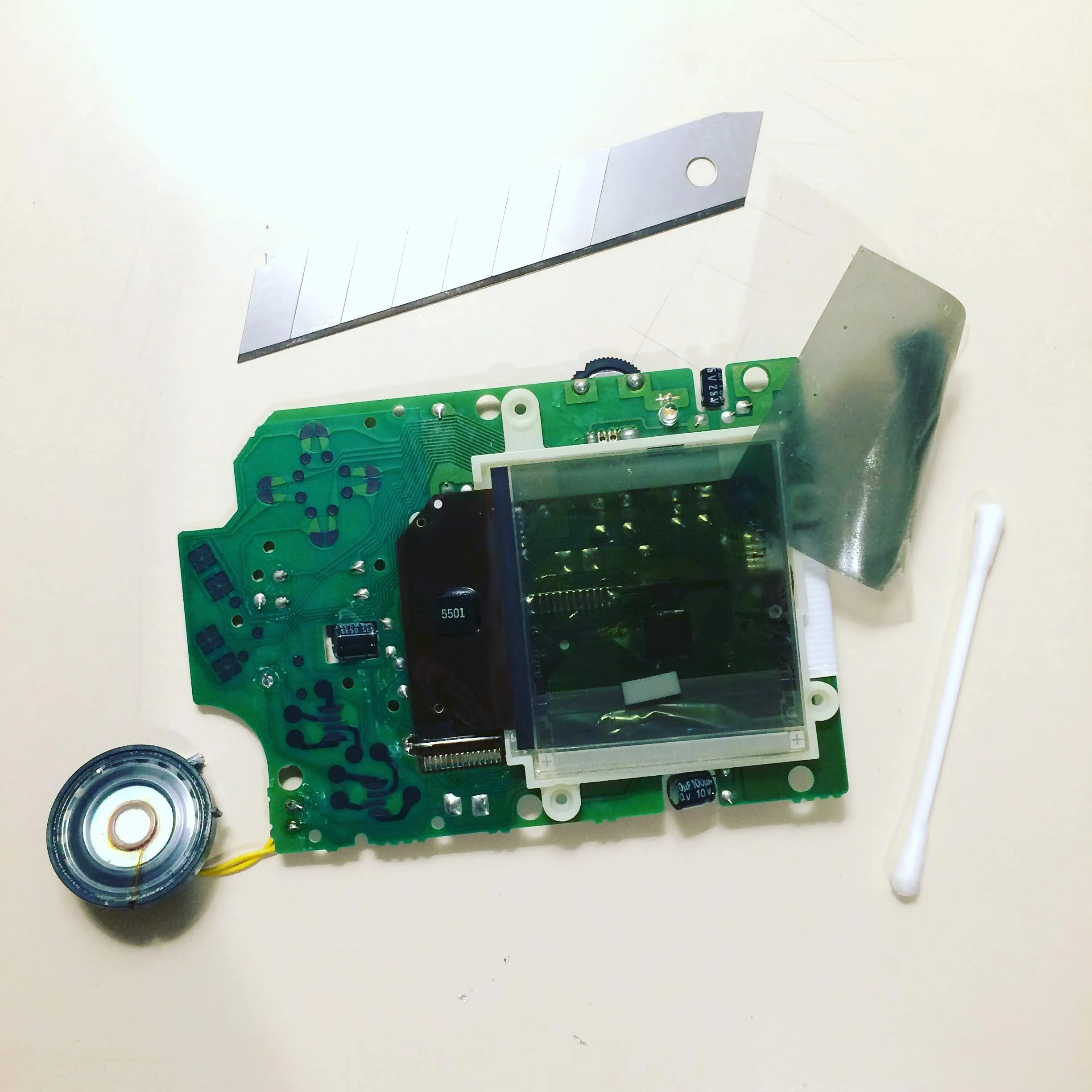 A Game Boy circuit board on a white desk with the LCD attached, the peeled reflective polarising film lying beside it as a curled grey sheet. A snapped-off craft knife blade and a cotton swab sit above and to the right, and the round speaker is detached at the lower left.