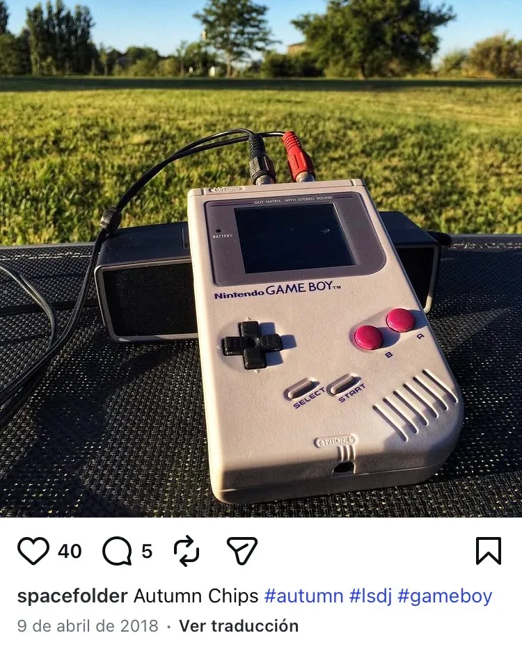 An assembled grey Game Boy propped on a dark table outdoors at golden hour, two RCA cables running from the top of it into a small black battery-powered speaker behind. Long grass and trees fill the background in low sun.
